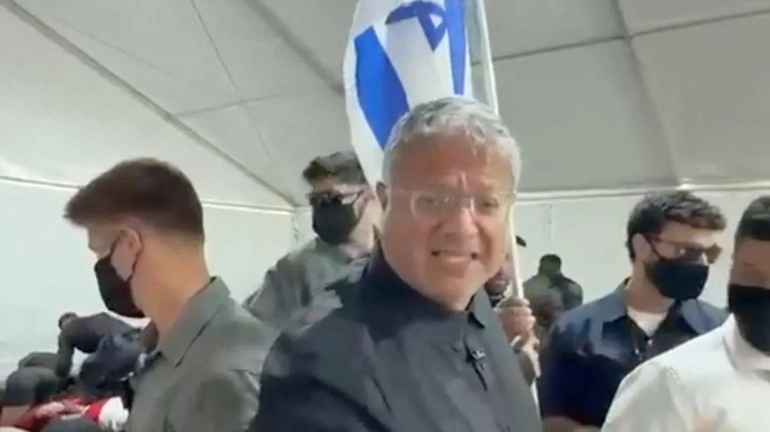 Reuters Itamar Ben Gvir standing holding an Israeli flag with several people standing around him in dark classes and masks 